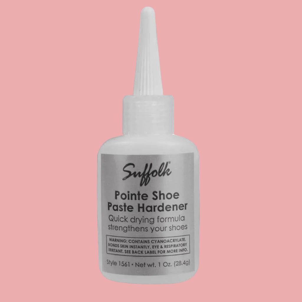 A bottle of Suffolk Pointe Shoe Paste Hardener with a white plastic applicator cap and a gray label, shown on a light pink background.